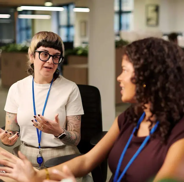 Two people sat side by side at a desk talking to each other