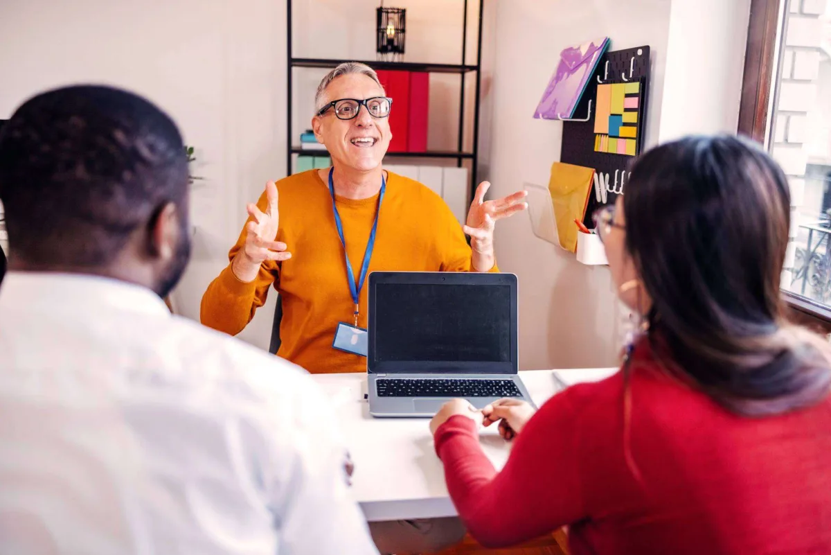 A man wearing a lanyard talks with open hands across a desk and laptop to two colleagues in a meeting